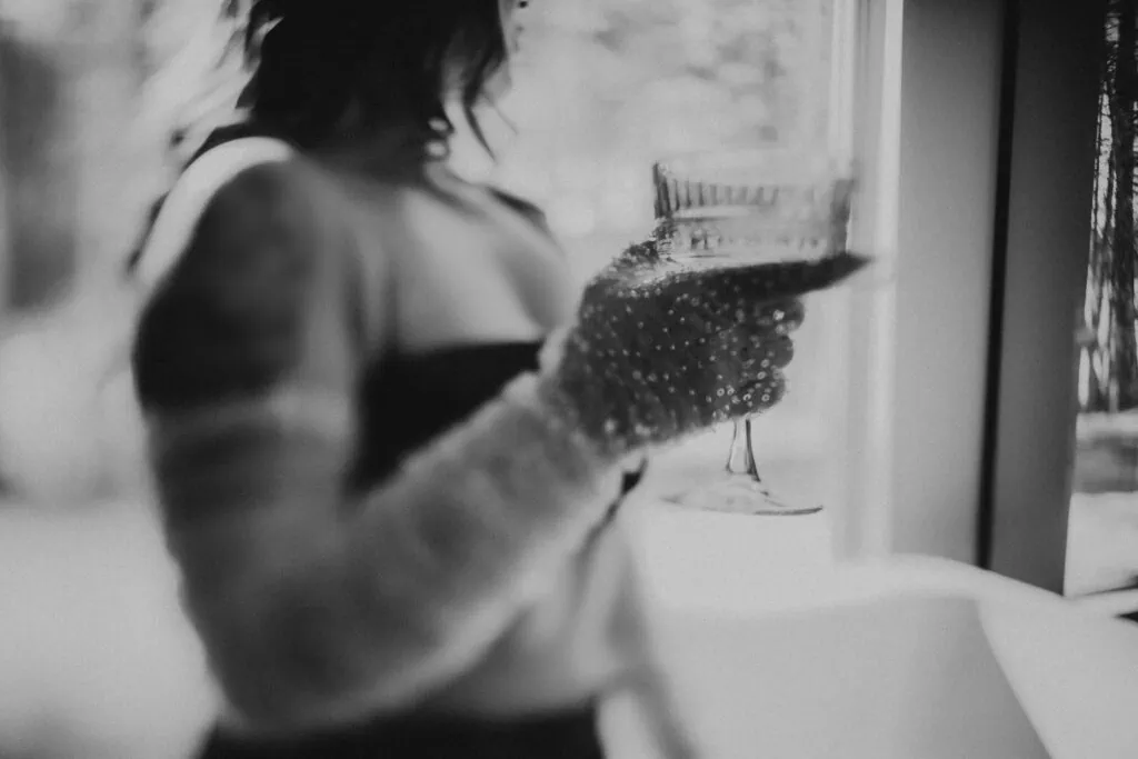 A black and white snapshot encapsulates a moment of grace featuring Ava Marie adorned in an intricately detailed sleeve, clutching a glittering glass near a window, radiating an air of thoughtful sophistication. Ava Marie Halifax's Elite Independent Companion